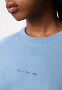 Sudadera azul claro con cuello acanalado. Presenta un logo en relieve de "Marc O'Polo DENIM" en el frente. Textura suave y diseño informal.