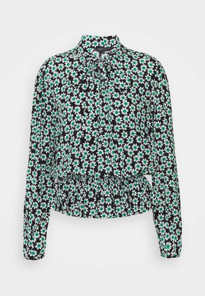 Blouse noire à manches longues avec imprimé marguerites blanches et vertes, col haut avec nœud noué et détail de taille froncée.