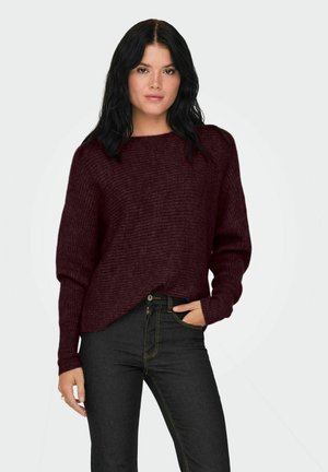 Pull en tricot bordeaux avec un col large, coupe ample et manches longues, associé à un jean foncé avec une couture contrastante.