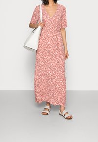 Sukienka maxi w kolorze koralowym z białym kwiatowym wzorem, głębokim dekoltem w kształcie V, krótkimi rękawami, marszczonym paskiem w talii, zestawiona z białą torebką i sandałami.