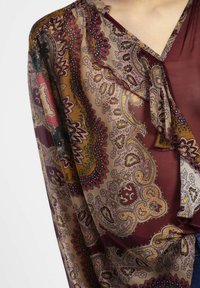 Blusa trasparente bordeaux con motivo paisley in tonalità di oro, verde e rosso; presenta un collo arricciato e maniche fluide.