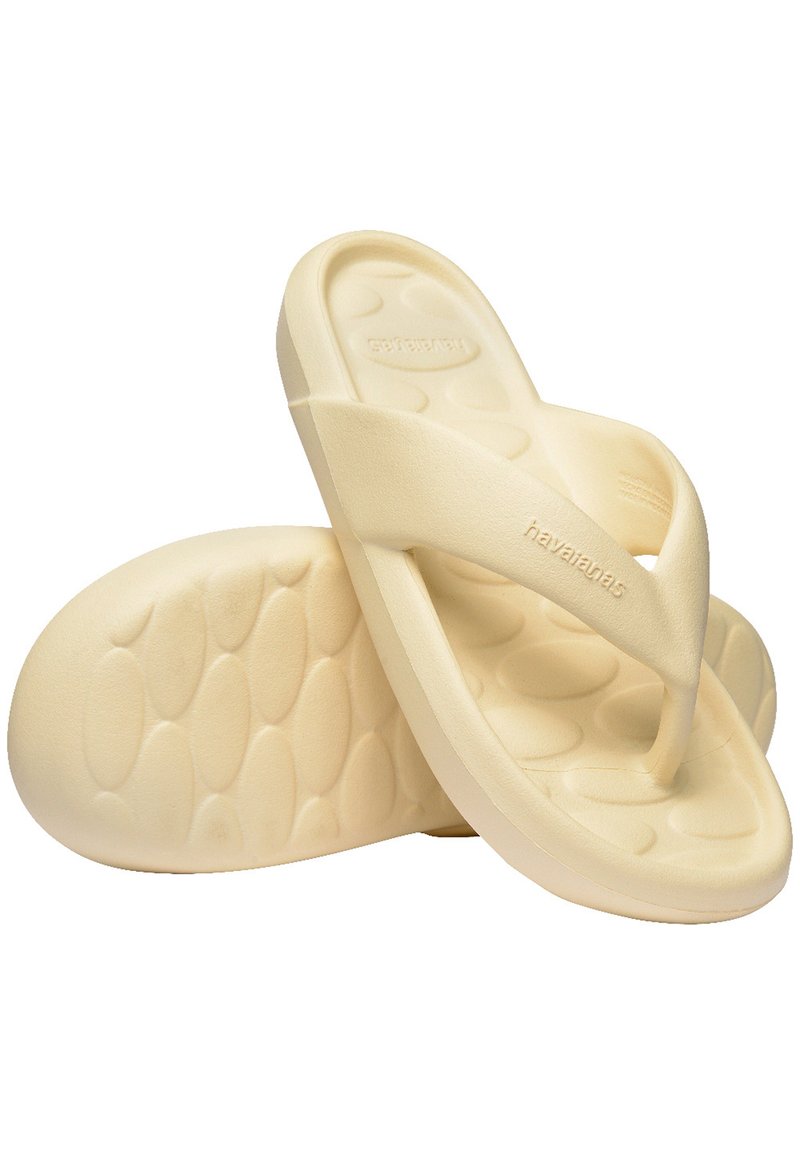 Béžové šľapky s textúrovanou podrážkou a hladkými, širokými popruhmi. Názov značky "Havaianas" je embosovaný na jednom popruhu.