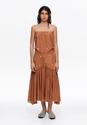 Joven mujer con vestido marrón sin tirantes con adornos de botones blancos y zapatillas grises abiertas en los dedos, de pie sobre un fondo blanco.