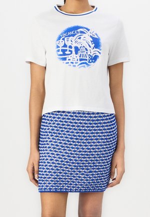 T-shirt blanc à manches courtes avec un motif bleu de palmier, verres à vin et texte « Vacances », associé à une jupe texturée bleue et blanche.