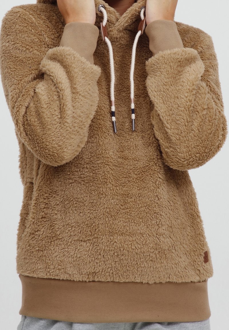 Personne portant un sweat à capuche beige en polaire douce avec des cordons blancs, tenant la capuche près du visage, visible de la poitrine au cou.