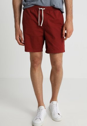 Homme portant un short décontracté rouge avec un cordon blanc et des baskets blanches, debout devant un fond clair uni.