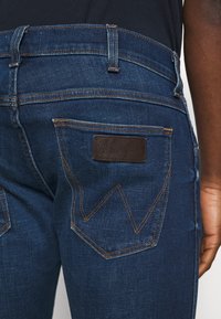 I jeans in denim blu scuro presentano un'etichetta in pelle e cuciture distintive sulle tasche posteriori, mettendo in risalto un design classico.