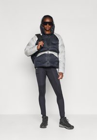 Sølv- og marineblå pufferjakke med hætte, der har vandrette striber. Bæres over sorte tætsiddende leggings og vandrestøvler. Modellen holder en rygsæk.