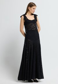 Sort blonde maxi kjole med en tætsiddende overdel, firkantet halsudskæring og skulderstropper med sløjfe. Har en flydende, lagdelt nederdel.