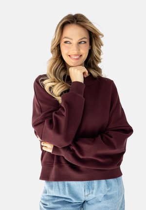 Młoda kobieta o długich blond włosach, ubrana w bordową bluzę i niebieskie jeansy, uśmiechająca się i opierająca podbródek na dłoni.