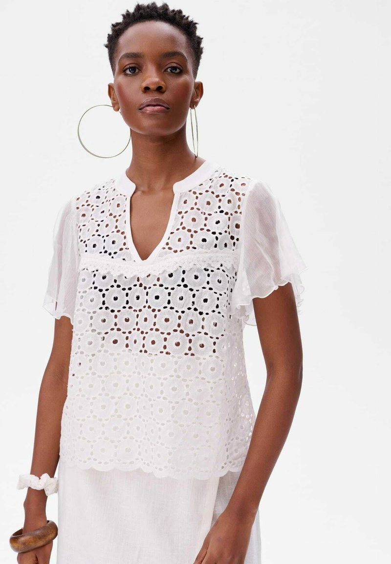 Biała bluzka z krótkim rękawem, ozdobiona haftem, z dekoltem w kształcie litery V i falbaną na dole. Wykonana z lekkiego materiału o teksturowanym, przezroczystym wykończeniu.
