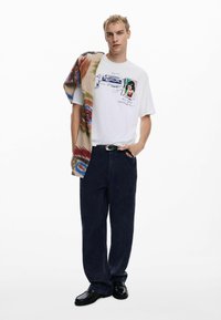 Valge T-särk trükitud graafikaga, tumedast sinisest laiadest pükstest ja mitmevärvilisest mustriga seljakattest, mis on visatud õlale. Mustad kingad.