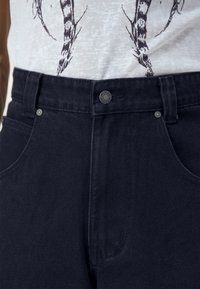 Tmavě modré džíny s vysokým pasem, kovovým knoflíkem a stříbrnými nýty. Obsahují dvě boční kapsy a viditelnou texturovanou látku.