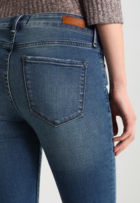 Jeans de mezclilla azul oscuro con una textura suave, que cuentan con un bolsillo trasero y un parche de cuero marrón con el logo en la pretina.
