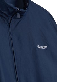 Granatowa kurtka z wysokim kołnierzem, zamek błyskawiczny i podszewka z siateczki. Posiada białe, haftowane logo po lewej stronie. Gładka tekstura materiału.