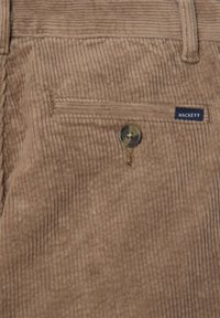 Béžové kordurové nohavice so zvislými rebrami, s gombíkovým zapínaním a malou modrou nálepkou s nápisom "HACKETT" na zadnej strane vrecka.