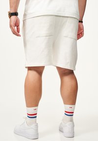 Weiße Baumwollshorts mit zwei hinteren Taschen, kombiniert mit weißen Sneakers und mehrfarbigen Socken mit gestreiften Muster in Rot, Blau und Weiß.
