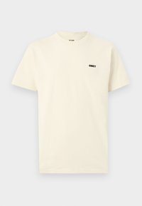 Μπλουζάκι κρεμ με κοντά μανίκια και στρογγυλή λαιμόκοψη, με μικρό μαύρο λογότυπο "OBEY" στο επάνω αριστερό μέρος του στήθους, μονόχρωμο φόντο.