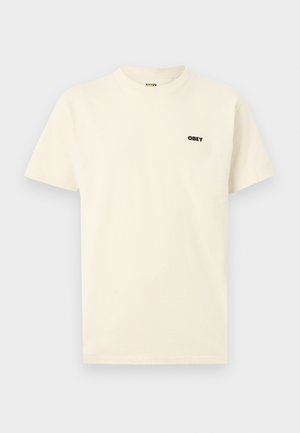 Krém színű, rövid ujjú, kerek nyakú póló kis fekete „OBEY” logóval a bal felső mellrészen, egyszínű háttérrel.