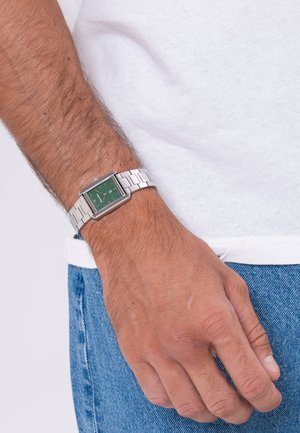 Mannsarm med en sølvfarget metallreim klokke med grønn kvadratisk urskive, nær en hvit skjorte og blå denimjeans.