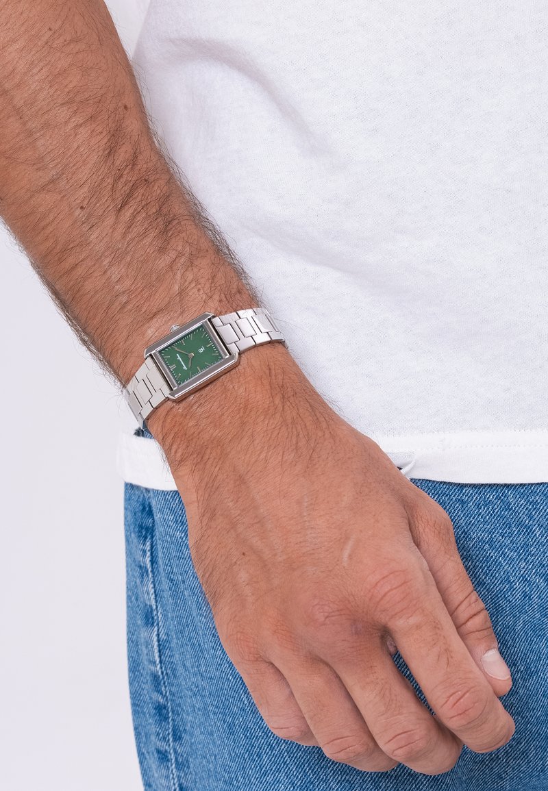 Ανδρικός καρπός που φορά ένα ασημί μεταλλικό ρολόι με πράσινο τετράγωνο καντράν, κοντά σε λευκό πουκάμισο και μπλε τζιν παντελόνι.