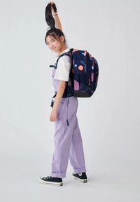Ragazza adolescente che indossa salopette viola, t-shirt bianca, sneakers nere e uno zaino colorato, mentre tiene su la coda di cavallo con una mano.