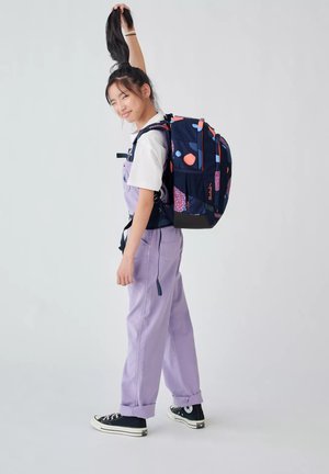 Fille adolescente portant une salopette lavande, un t-shirt blanc, des baskets noires et un sac à dos coloré, tenant sa queue de cheval avec une main.