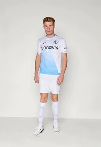 Vyras, vilkintis šviesiai mėlyną ir baltą futbolo uniformą, su baltomis trumpomis kelnėmis, ilgomis baltomis kojinėmis ir baltomis sportinėmis bateliais, stovi prieš paprastą foną.