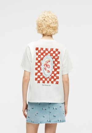 Osoba o kręconych blond włosach, ubrana w białą koszulkę z czerwonym wzorem w kratę, trzymająca talerz z truskawkami, w połączeniu z niebieską, kwiecistą spódnicą dżinsową.