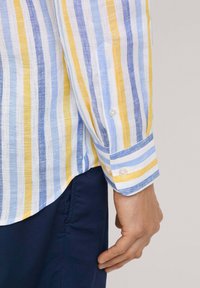 Camisa a rayas en lino de color azul claro, amarillo y blanco; presenta un puño abotonado y un dobladillo curvo. Se lleva con pantalones azul marino oscuros.