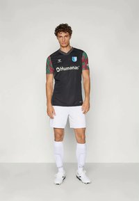 Black football jersey with patterned short sleeves, featuring a logo and "Humanas" text. Worn with white shorts and socks, complemented by white cleats.