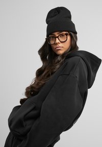 Hoodie noir surdimensionné avec cordon de serrage et poche avant ; assorti à un bonnet en tricot noir et de grandes lunettes rectangulaires en écaille de tortue.