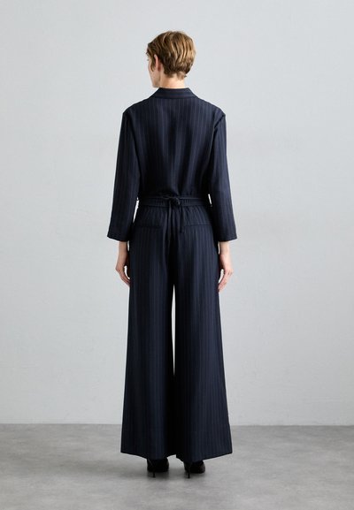 Mono de pierna ancha en azul marino con rayas, que presenta un lazo envolvente en la cintura, mangas largas y dos bolsillos traseros. Textura de tejido suave.