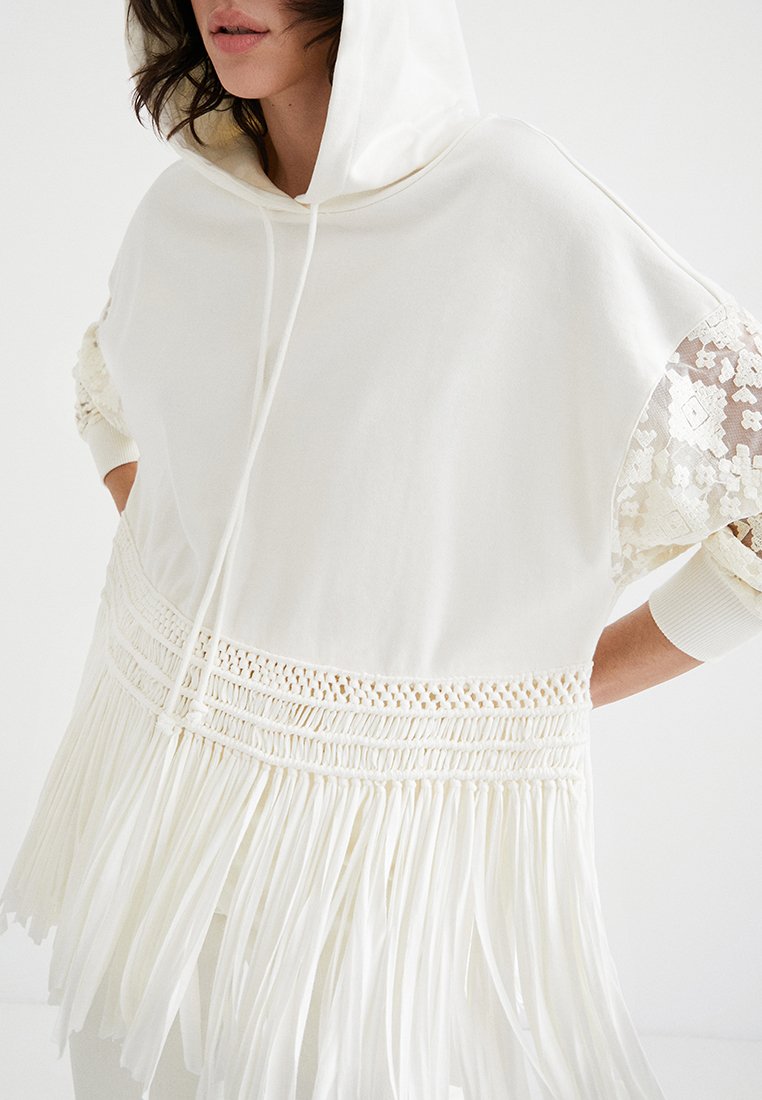 Donna che indossa un top bianco con cappuccio, maniche in pizzo e dettagli in frange intorno alla vita, su uno sfondo bianco semplice.