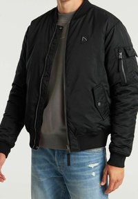 Man draagt een zwarte bomberjack met rits- en drukknoopzakken over een grijze sweatshirt en lichtblauwe gescheurde spijkerbroek.