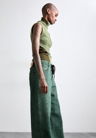 Πράσινο αμάνικο πλεκτό τοπ με ψηλό γιακά και ριγέ στρίφωμα, συνδυασμένο με φαρδιά πράσινα τζιν παντελόνια με κορδόνι στη μέση και πλαινές τσέπες.