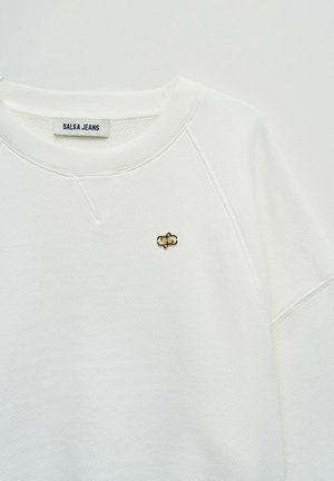 Sudadera blanca con textura, cuello redondo, pequeño pin de logo metálico dorado y negro en el pecho, y etiqueta interna que dice "SALSA JEANS".