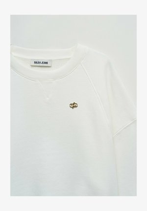 Sudadera blanca con textura, cuello redondo, pequeño pin de logo metálico dorado y negro en el pecho, y etiqueta interna que dice "SALSA JEANS".