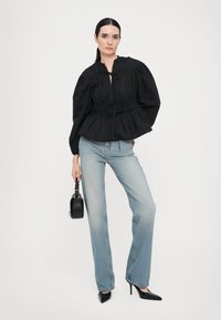 Svart bluse i stoff med puffskuldre og smocket midje; sammen med lyseblå jeans og spisse svarte hæler; holder en liten svart håndveske.