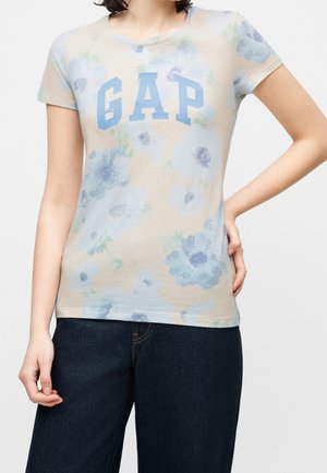 Personne portant un t-shirt beige à manches courtes à motif floral avec le logo bleu "GAP" et un jean bleu foncé, debout avec une main sur la hanche et l'autre détendue.