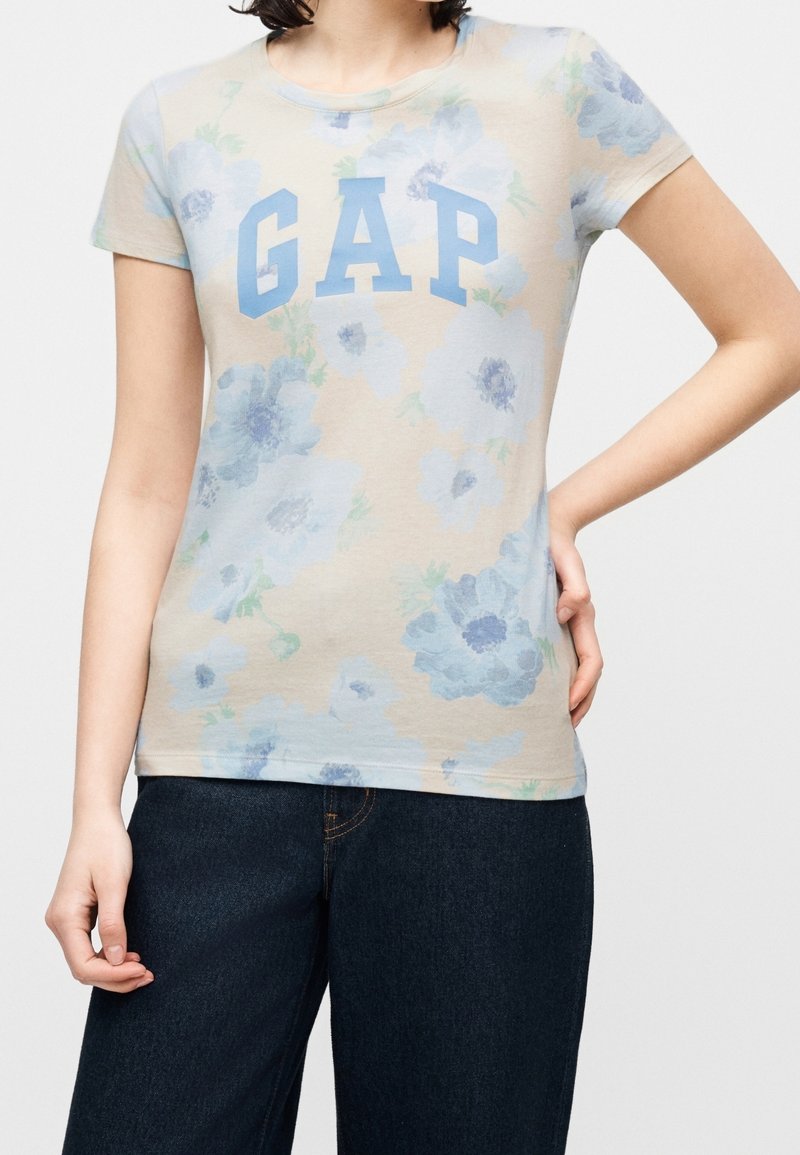 Personne portant un t-shirt beige à manches courtes à motif floral avec le logo bleu "GAP" et un jean bleu foncé, debout avec une main sur la hanche et l'autre détendue.