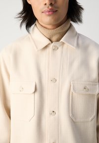 Light beige textured shirt jacket with a collared neckline, two front pockets, and button closures. Worn over a tan turtleneck.