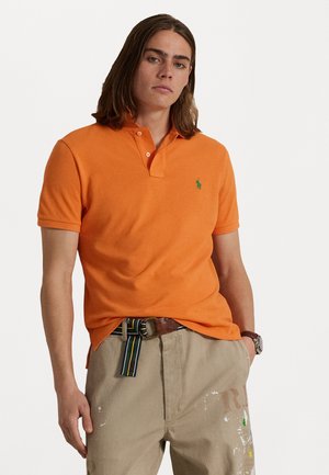 Oranje polo shirt met een twee-knopen knoopsluiting en groen logo. Gecombineerd met beige broek en een meerkleurige gestreepte riem met verfspatten.