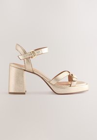 Gold metallic block-heeled sandal with ankle and toe straps, featuring two gold buckle closures on a neutral background.
