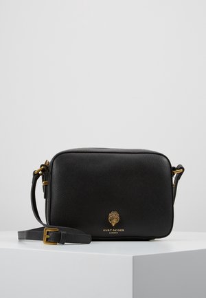 Sac bandoulière rectangulaire en cuir noir avec boucle dorée et logo, posé sur un piédestal blanc sur un fond gris clair uni.