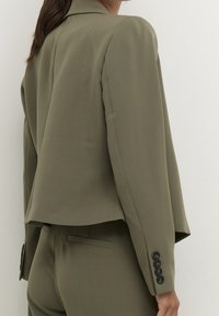 Olivově zelený ušitý blazer s mírně zkráceným střihem, klopovými límci a knoflíkovými manžetami. Hladká tkanina s minimalistickým designem.