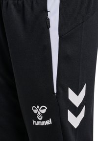Schwarze Sportpants mit weißen Seitenstreifen, ausgestattet mit einer Reißverschlusstasche und dem Hummel-Logo am unteren Bein, aus strukturiertem Material.