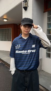 Marineblauw gestreept poloshirt met witte accenten, met de tekst "Santa Cruz" en sterren, gedragen over een wit shirt met lange mouwen. Zwarte pet.