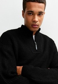 Czarny sweter z dzianiny z półzamkiem, prążkowaną fakturą i dekoltem w kształcie litery V. Materiał wydaje się gruby i ciepły, idealny do warstwowania.
