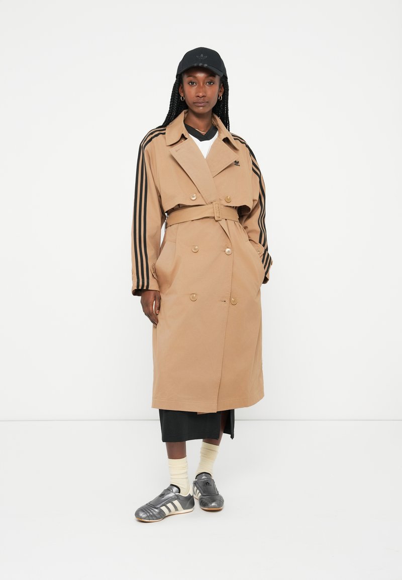 Tan dobbeltbrystet trench coat med sorte stribede ærmer, bælte i taljen og gyldne knapper. Sammen med en sort cap og grå sportsko.